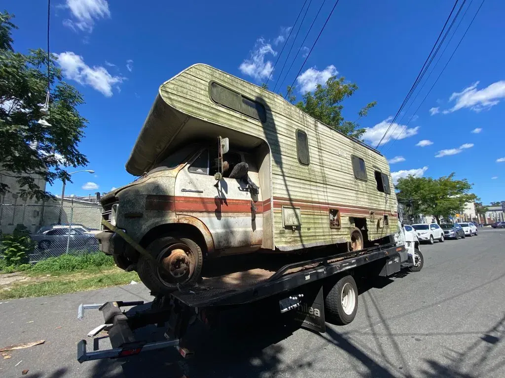 Old, weathered RV on a tow truck, being transported on a sunny, blue sky day.