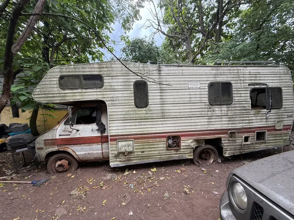 Old, weathered RV, parked outdoors, surrounded by trees and mud. Wheels flat, discolored paint, appears abandoned.
