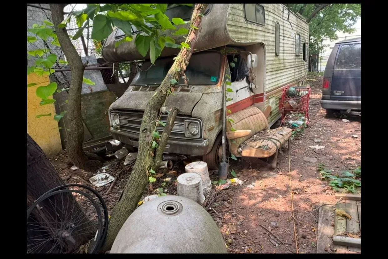 An old, weathered RV parked amongst trees and debris; the vehicle is run-down with visible rust and damage.