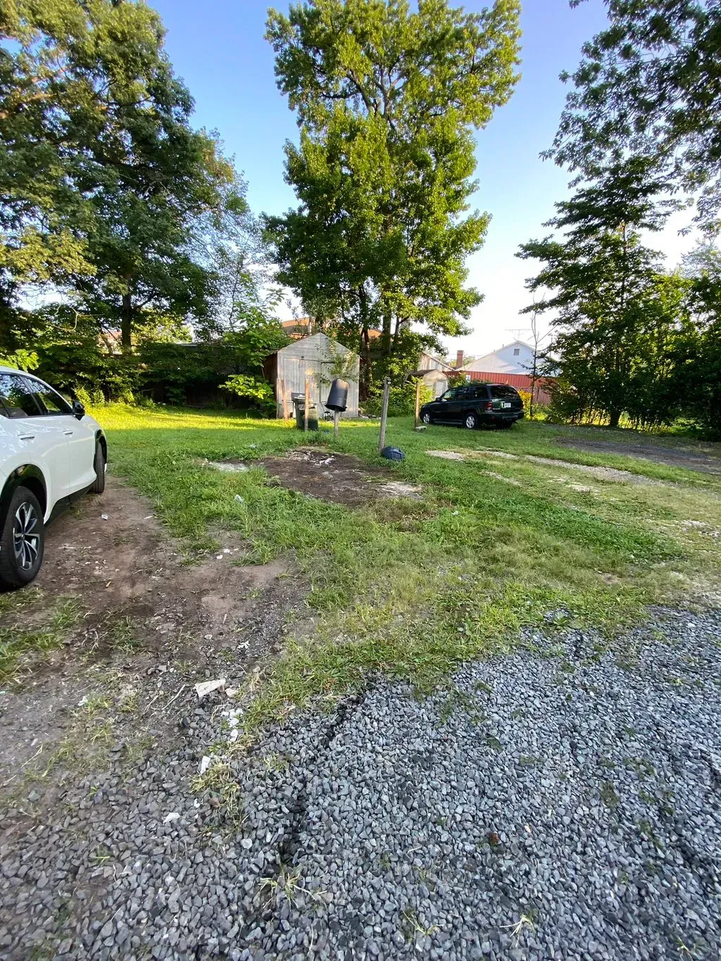 Gravel driveway with two cars parked on grass. Trees and a small building in the background under a blue sky.