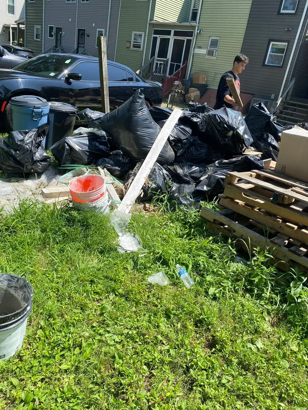 Pile of black trash bags with debris and a person sorting outdoors. Grassy area, sunlight.
