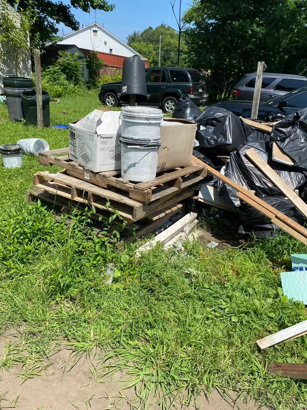 Pile of trash, including buckets, boxes, and bags on pallets, in a grassy yard; cars and house in the background.