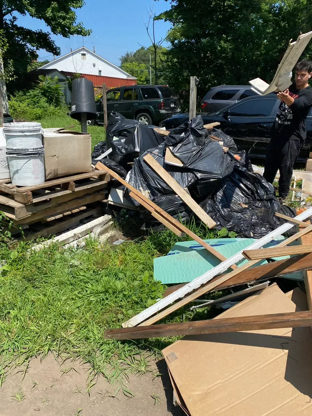 A person near a pile of trash bags, wooden planks, and building materials on a grassy lawn in front of a house.