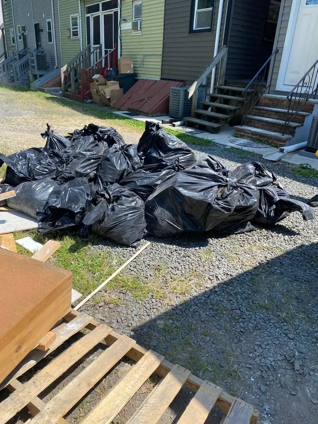 Pile of black trash bags on gravel near a building. Wooden pallet and cardboard boxes are in foreground.