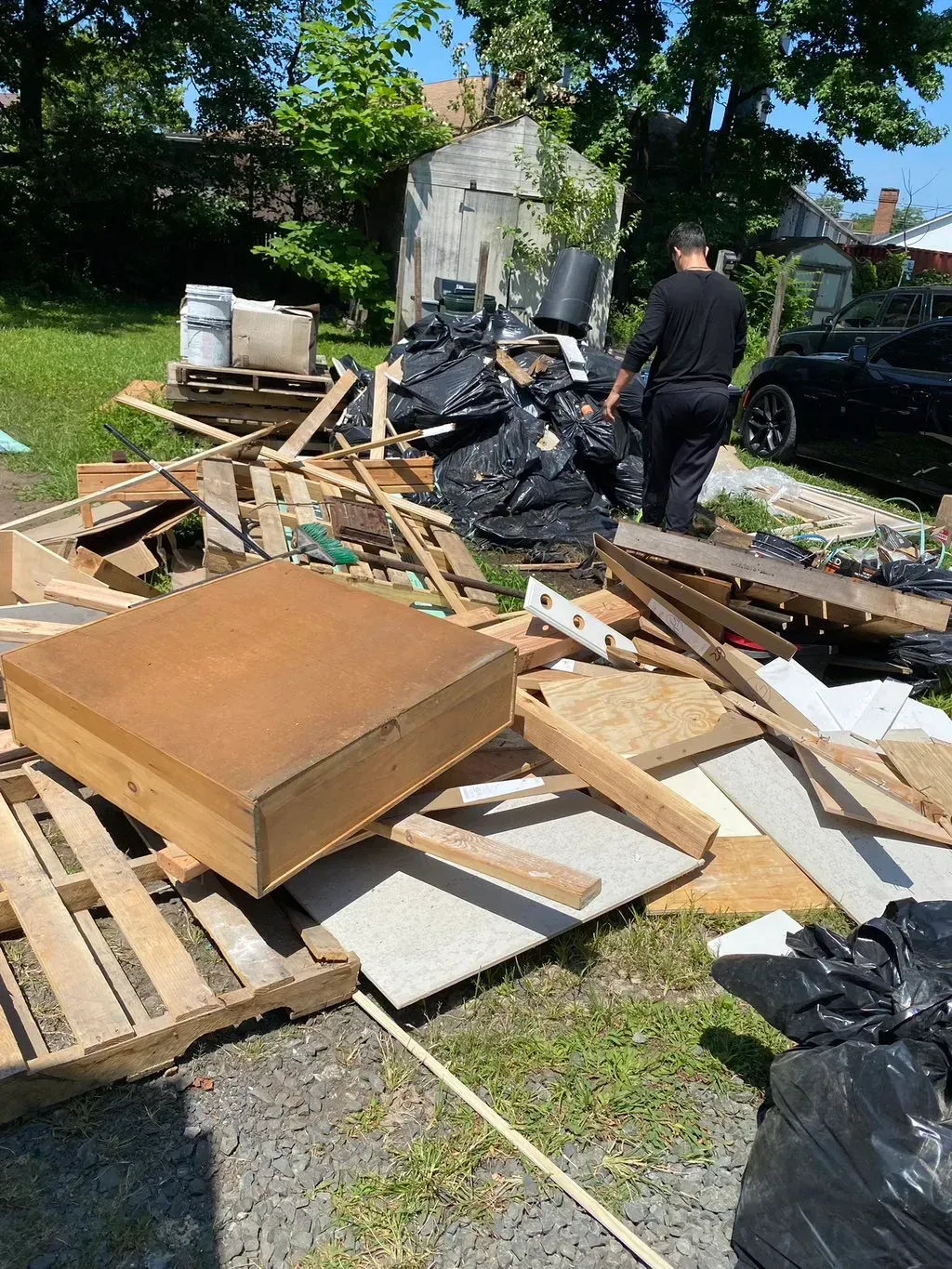 A man stands near a pile of debris including wood, black bags, and pallets in a backyard.