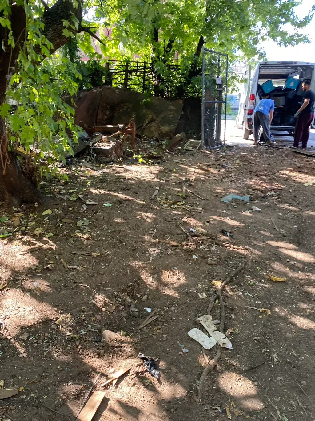 Dirt patch with debris, trees, and a fence. People unload items from a van.
