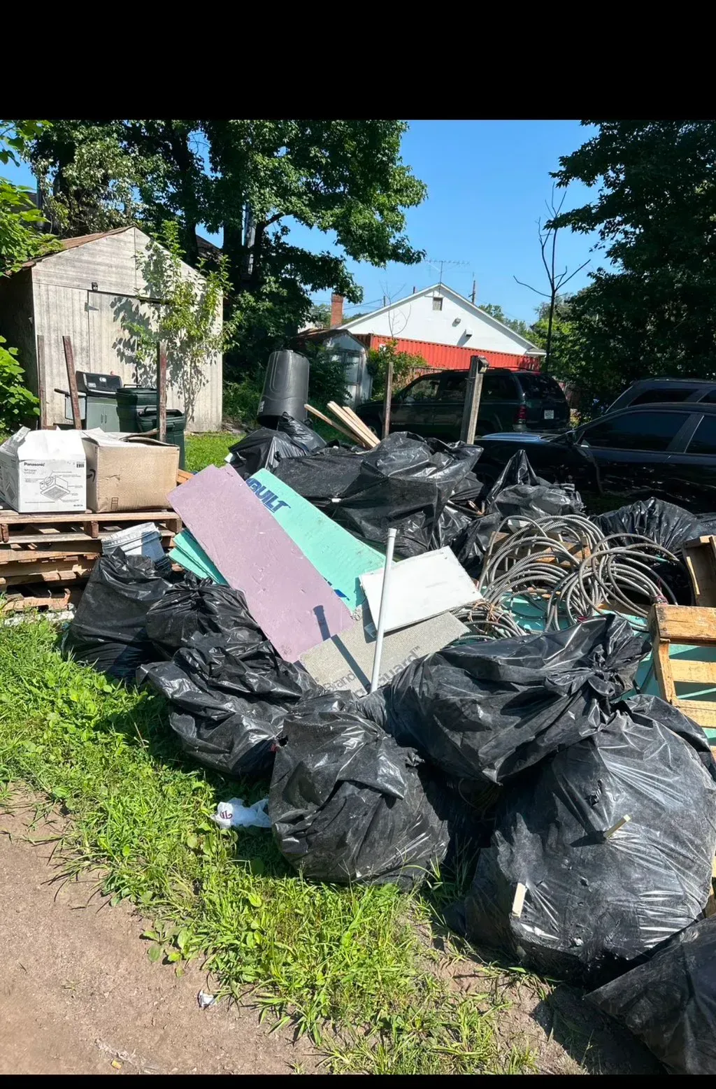 Pile of trash bags, debris, and construction materials in an outdoor setting near a shed and houses.
