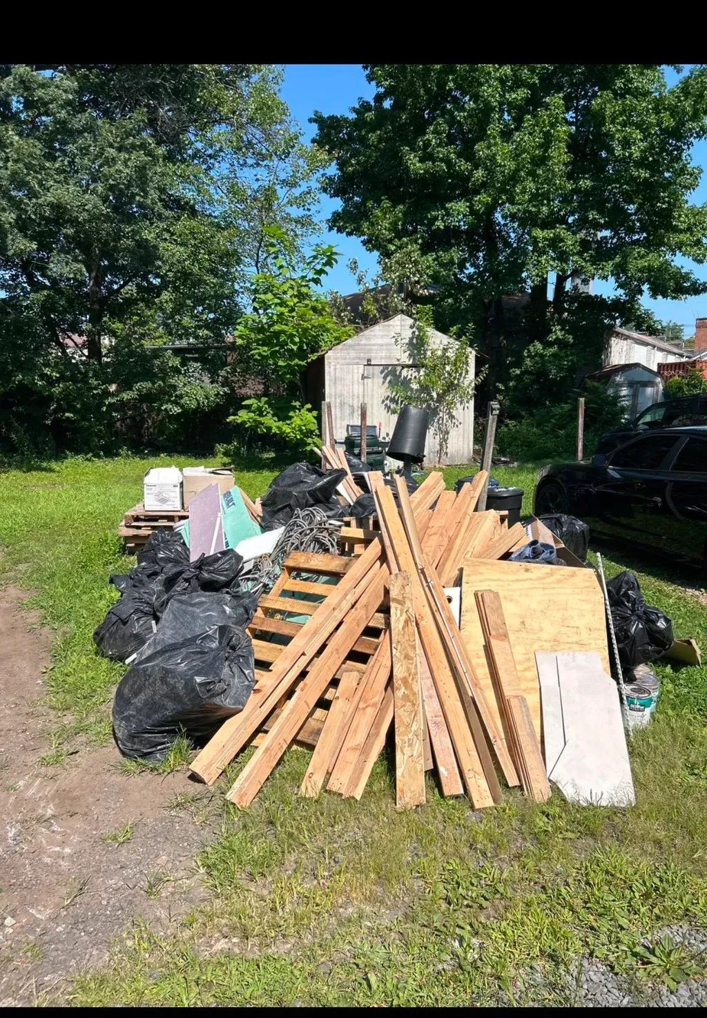 Pile of debris including wood, bags, and a pallet sits in front of a small shed in a grassy yard.