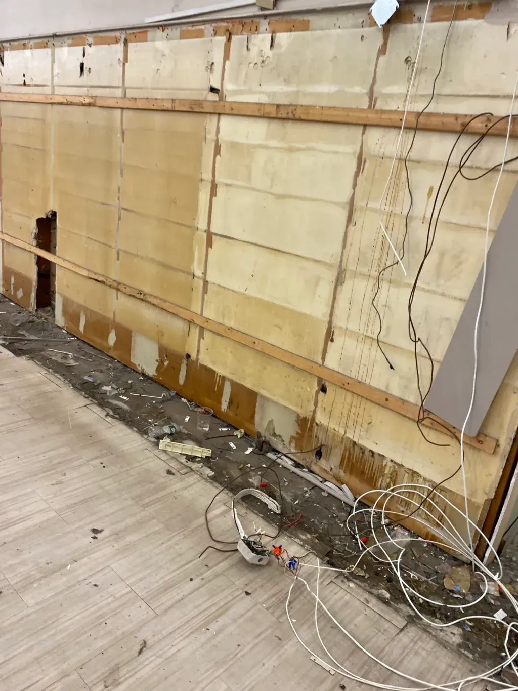 Exposed wall under renovation, showing wood paneling, wiring, debris, and flooring.