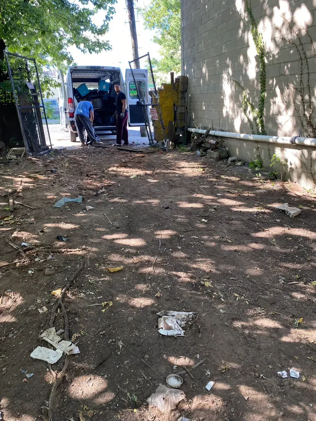 People loading a van on a dirt path, next to a building. Littered ground. Sunny day.