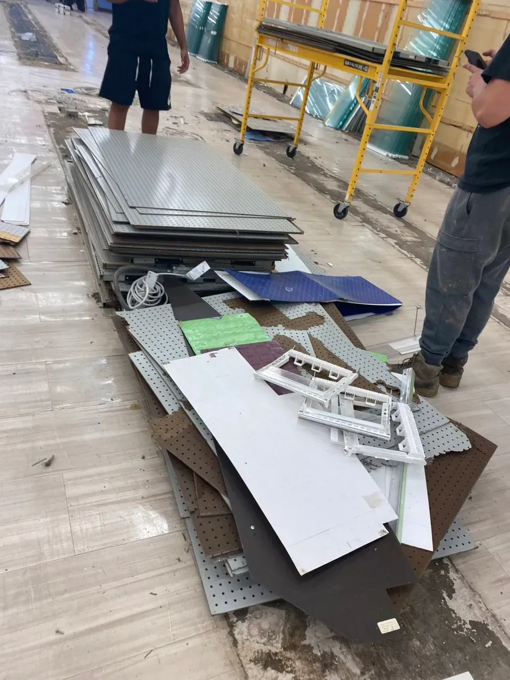 Pile of construction materials on a floor, with two people nearby and a yellow scaffold.