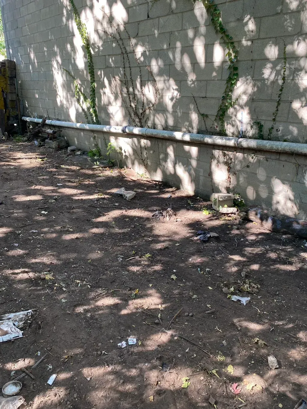 Dirt ground next to a concrete wall with a pipe. Shadows and some trash are visible.