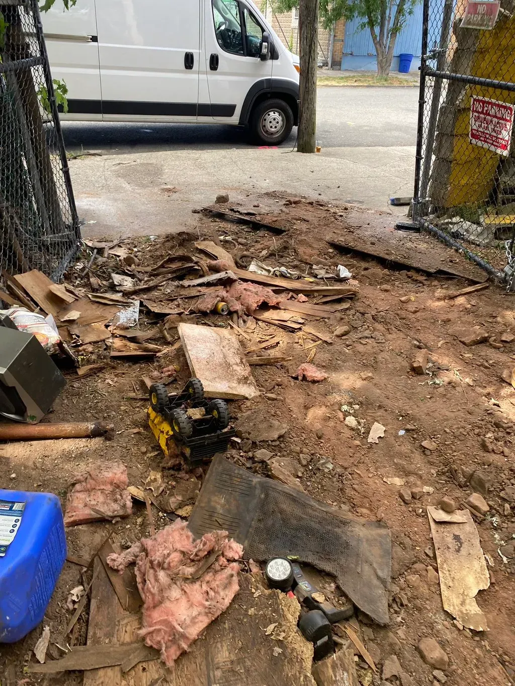 Debris-filled alleyway. White van parked. Litter including insulation, wood, and containers on muddy ground.