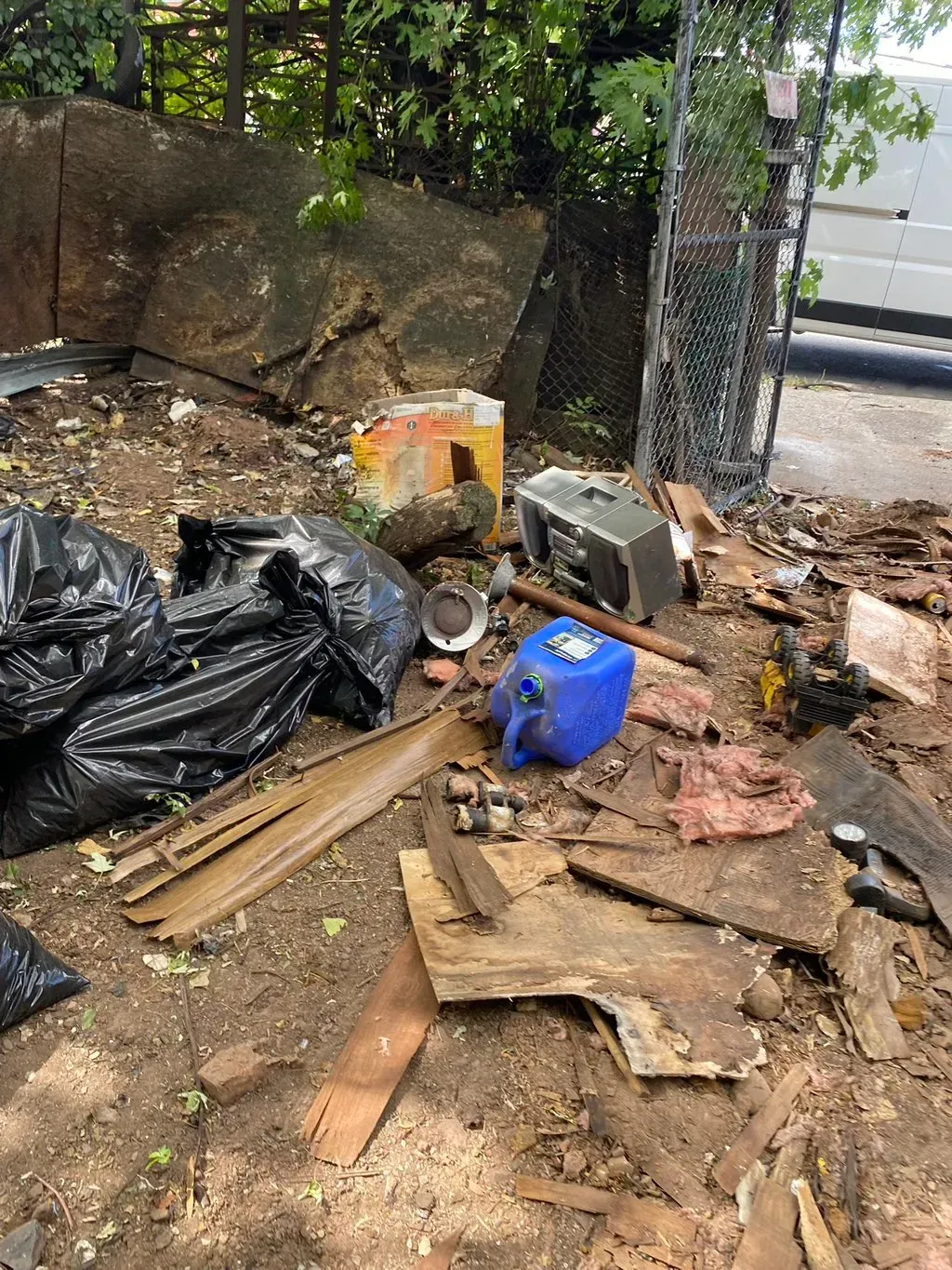 Pile of trash and debris in an outdoor area, including black bags, wood, a blue jug, and a radio.