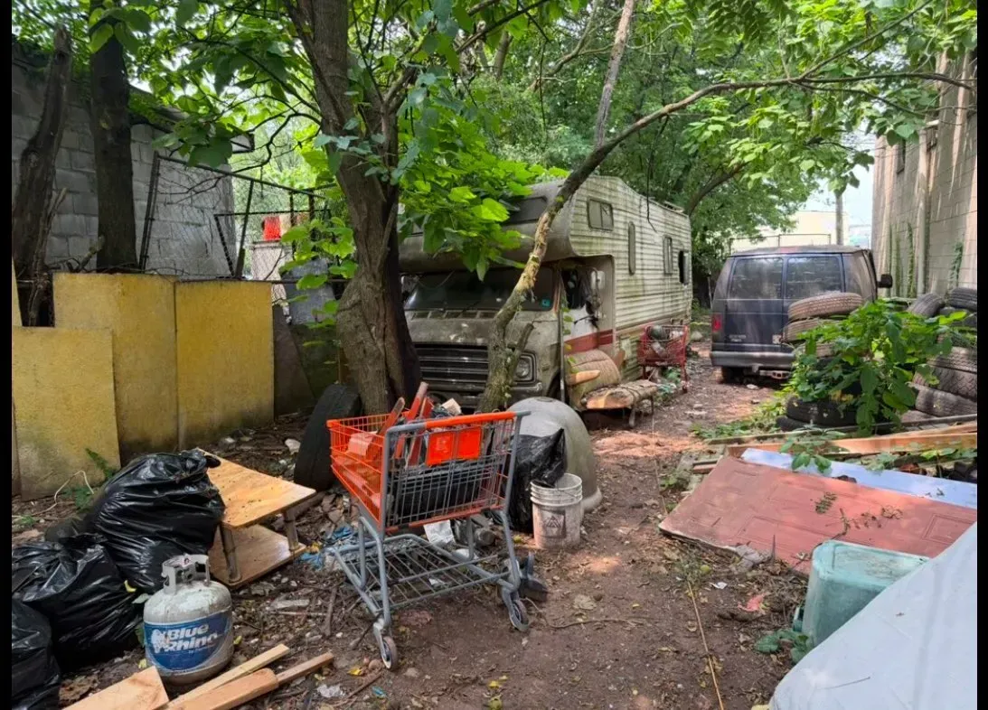 Overgrown alleyway with a trailer, trash, shopping cart, and debris; looks neglected.