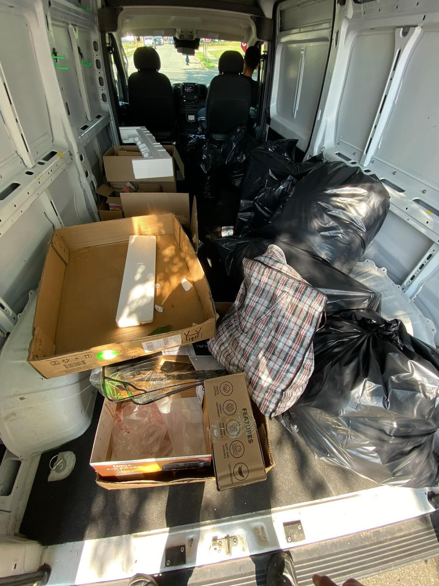 Interior of a cargo van filled with boxes, black bags, and other items. Two people are visible in the driver and passenger seats.