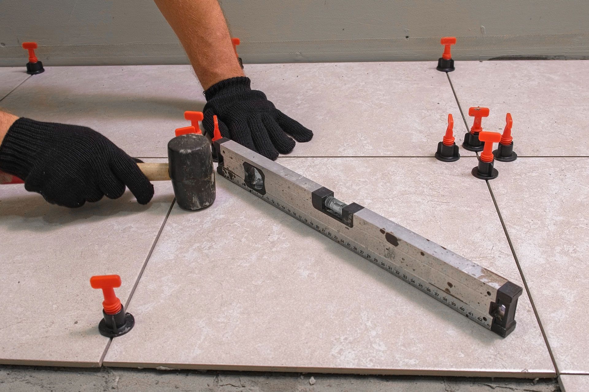 Gloved hands use a level and mallet to align floor tiles with suction cup spacers and clips