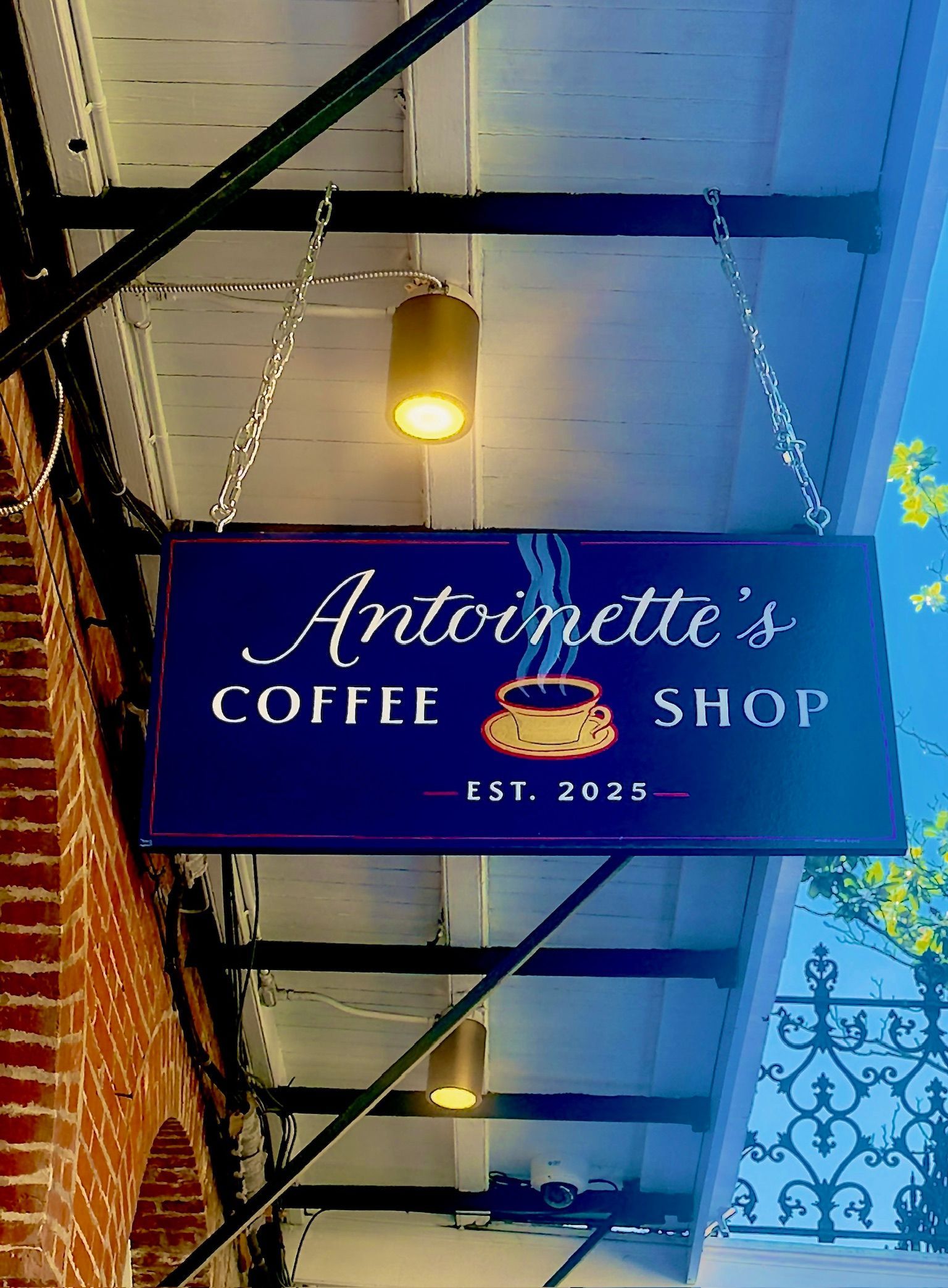 Sign for Antoinette's Coffee Shop, blue with white lettering, hanging under awning, brick wall to the left.