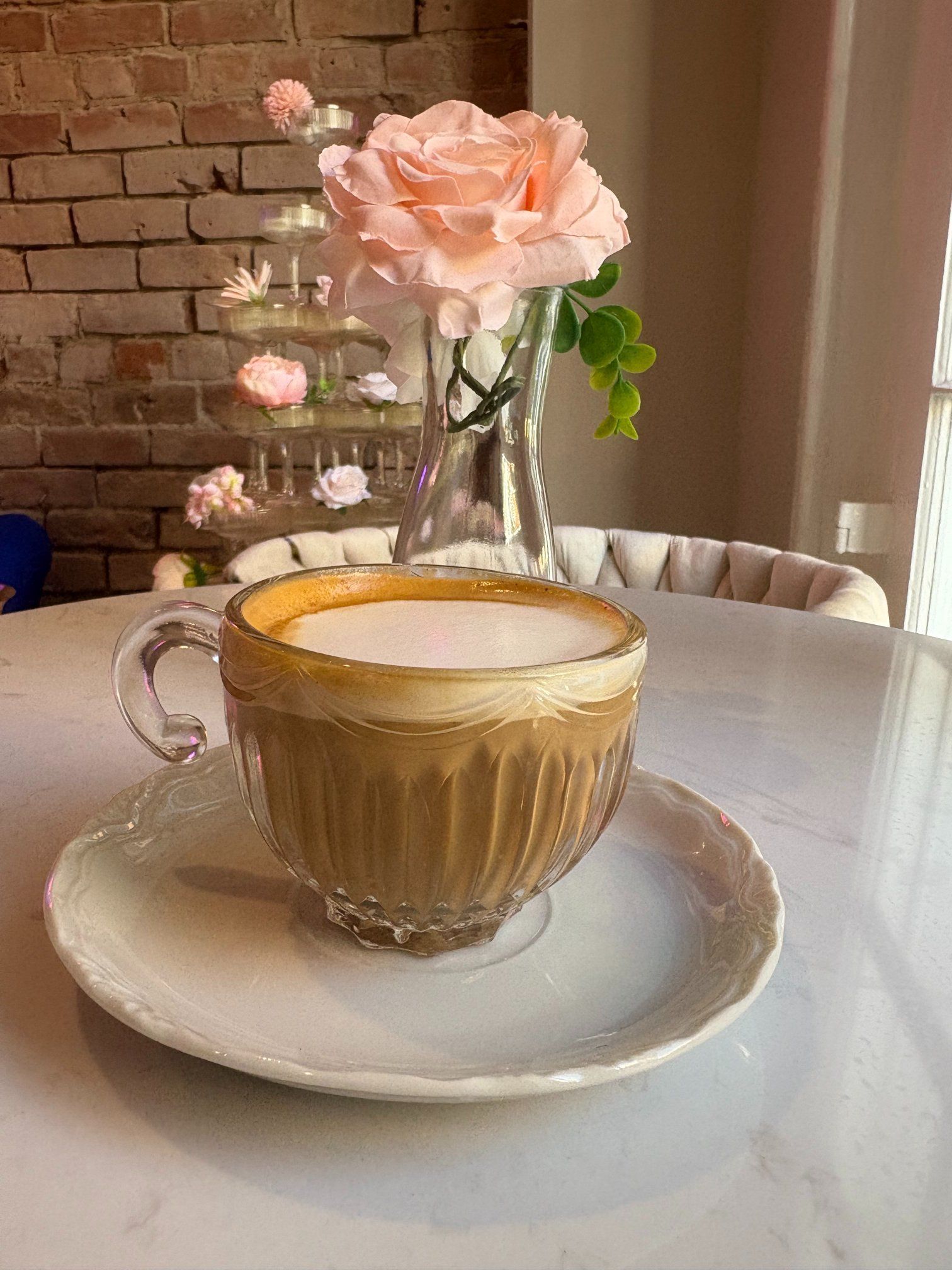 Cup of coffee on a saucer, near a vase with pink roses and a brick wall background.