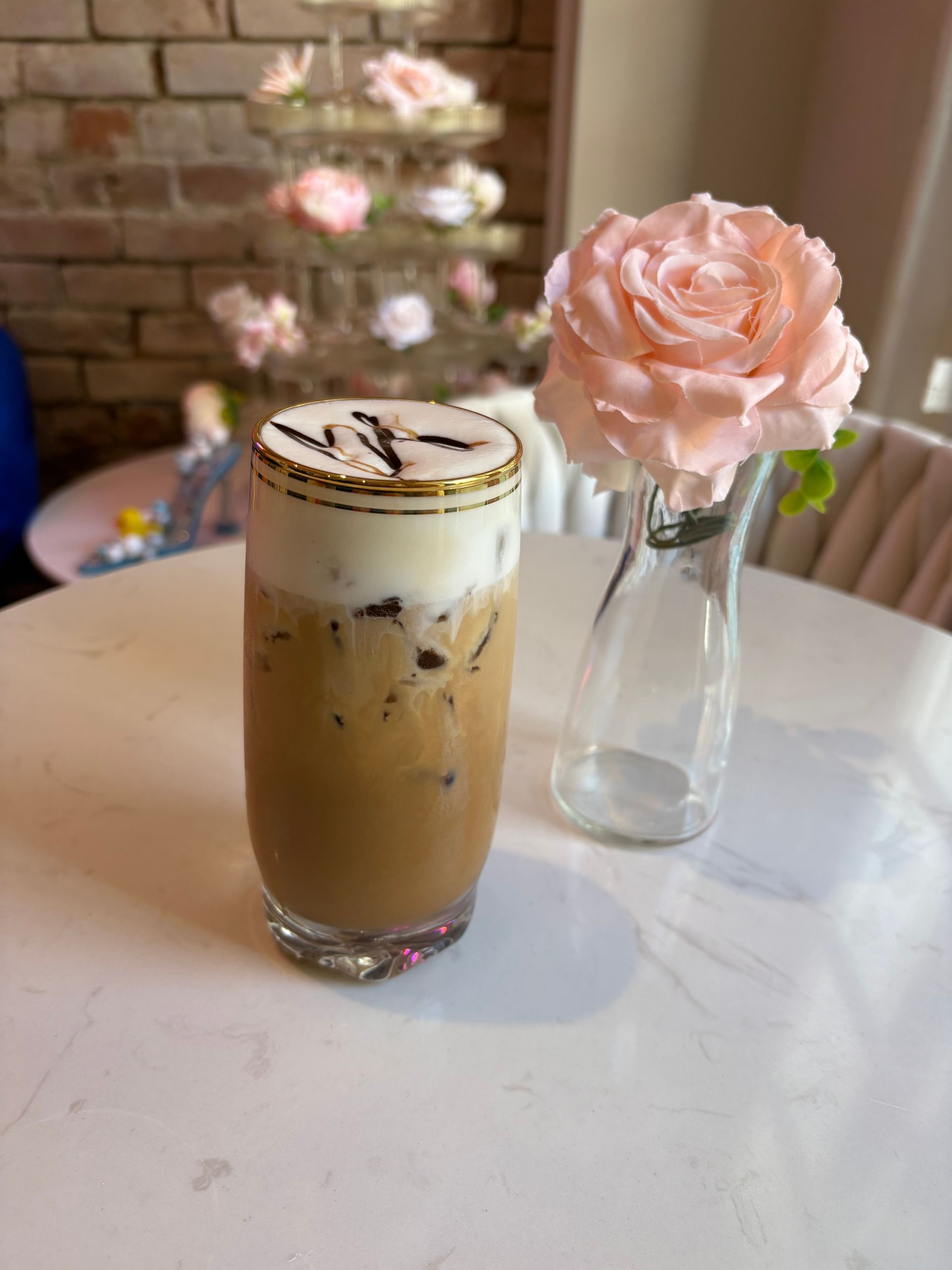 Iced coffee drink with foam and chocolate drizzle next to a vase with a pink rose.