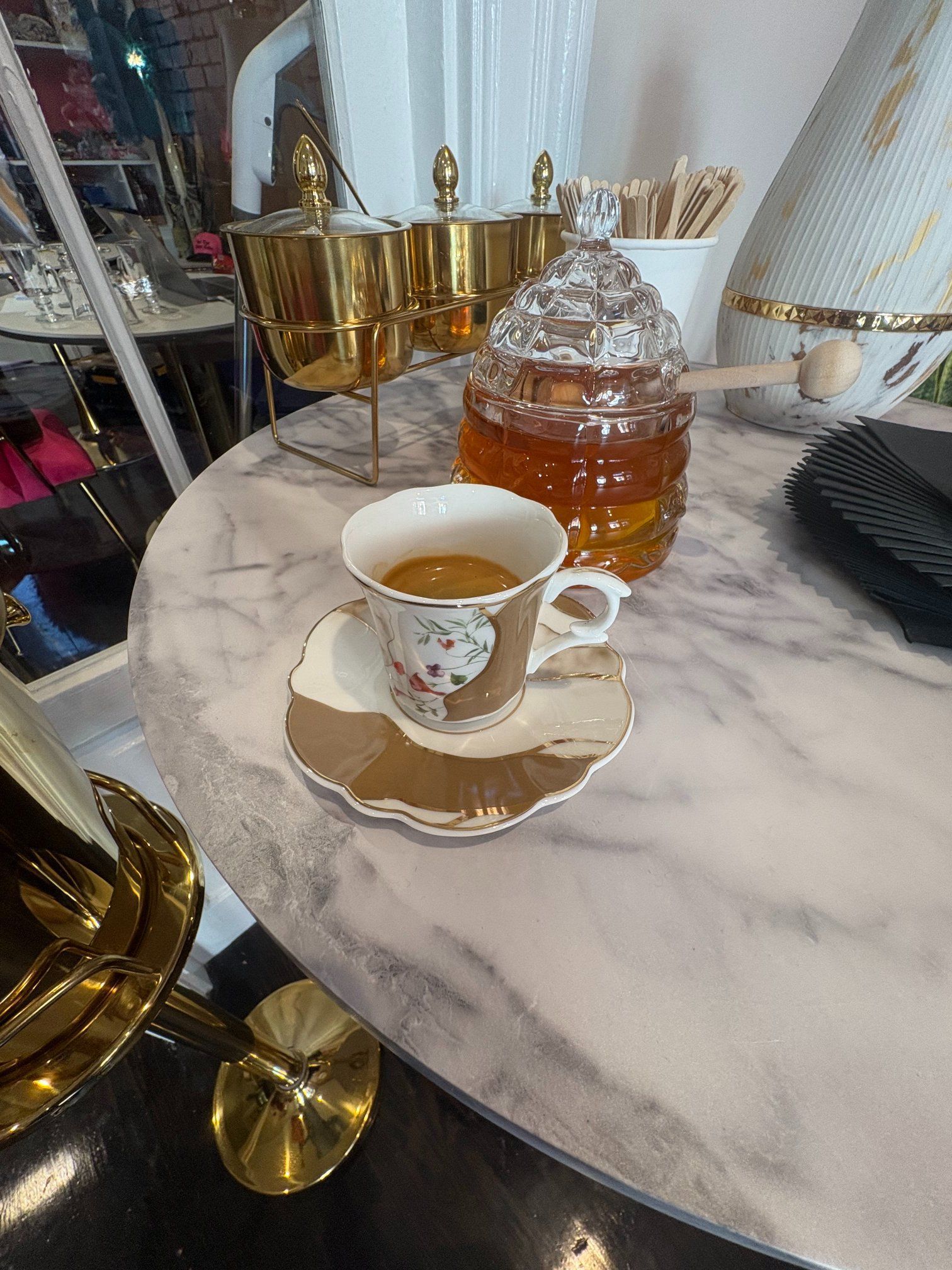 Cup of coffee on a saucer, jar of honey, and gold serving dishes on a marble table.