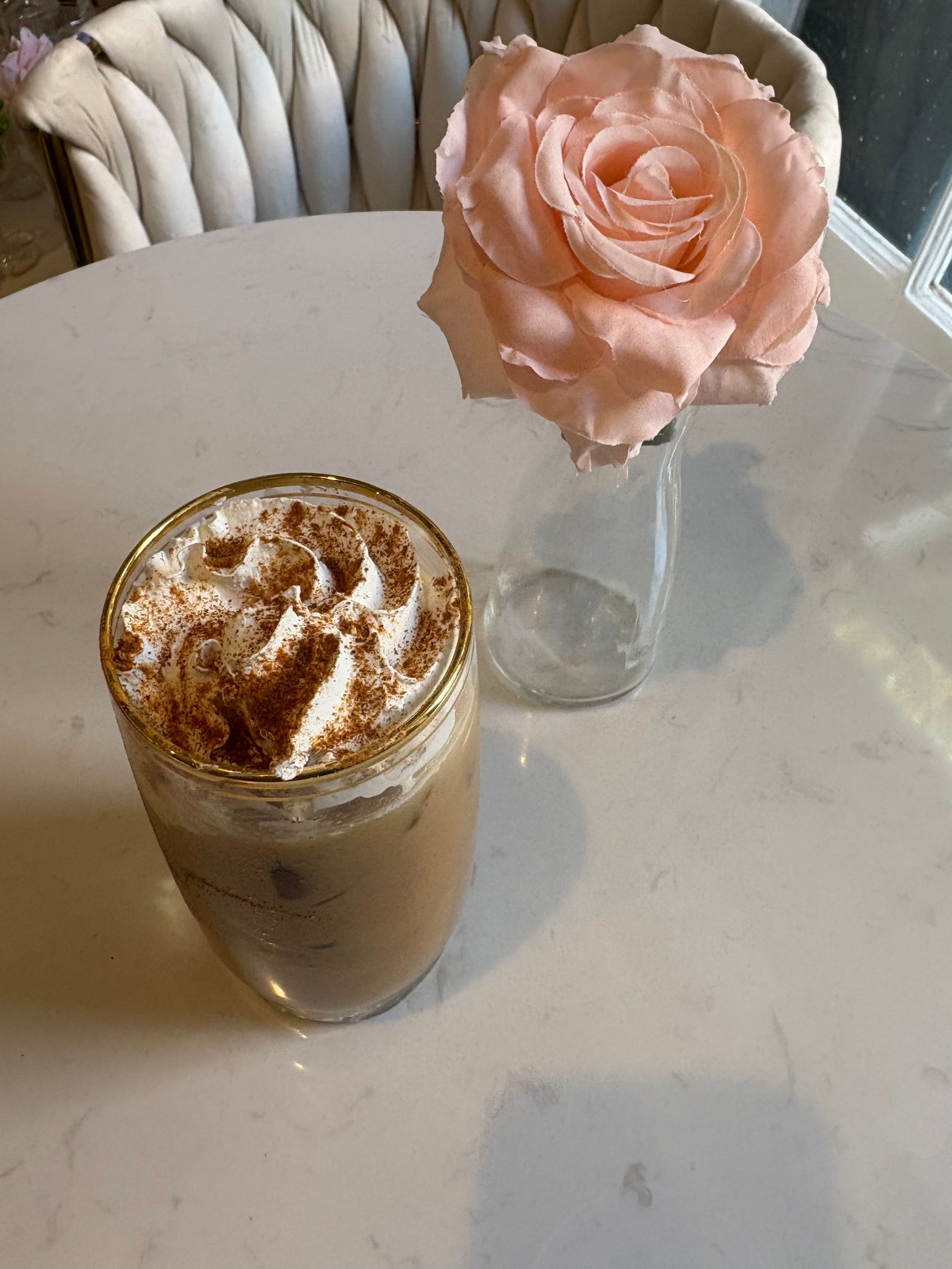 Iced coffee with whipped cream and cinnamon, beside a pink rose in a vase on a white table.