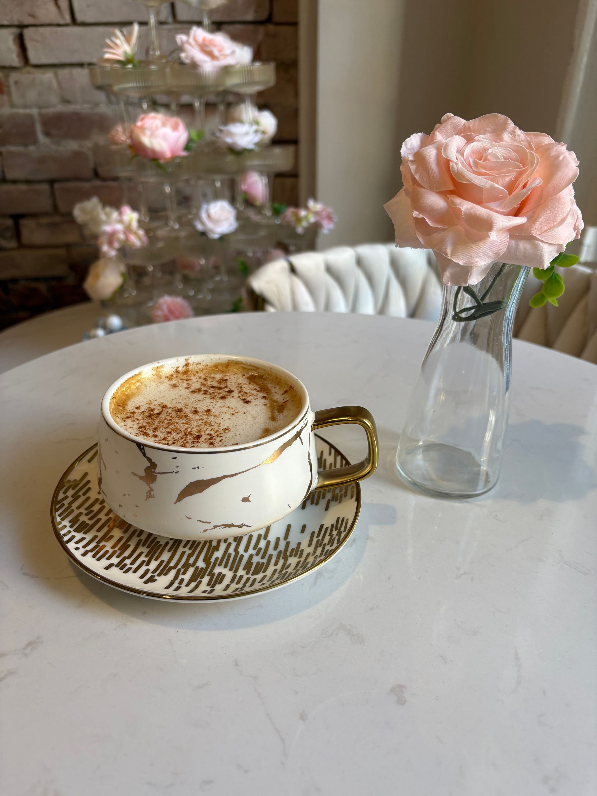 Coffee in a decorative cup on a white table next to a vase with a pink rose, with flowers in the background.