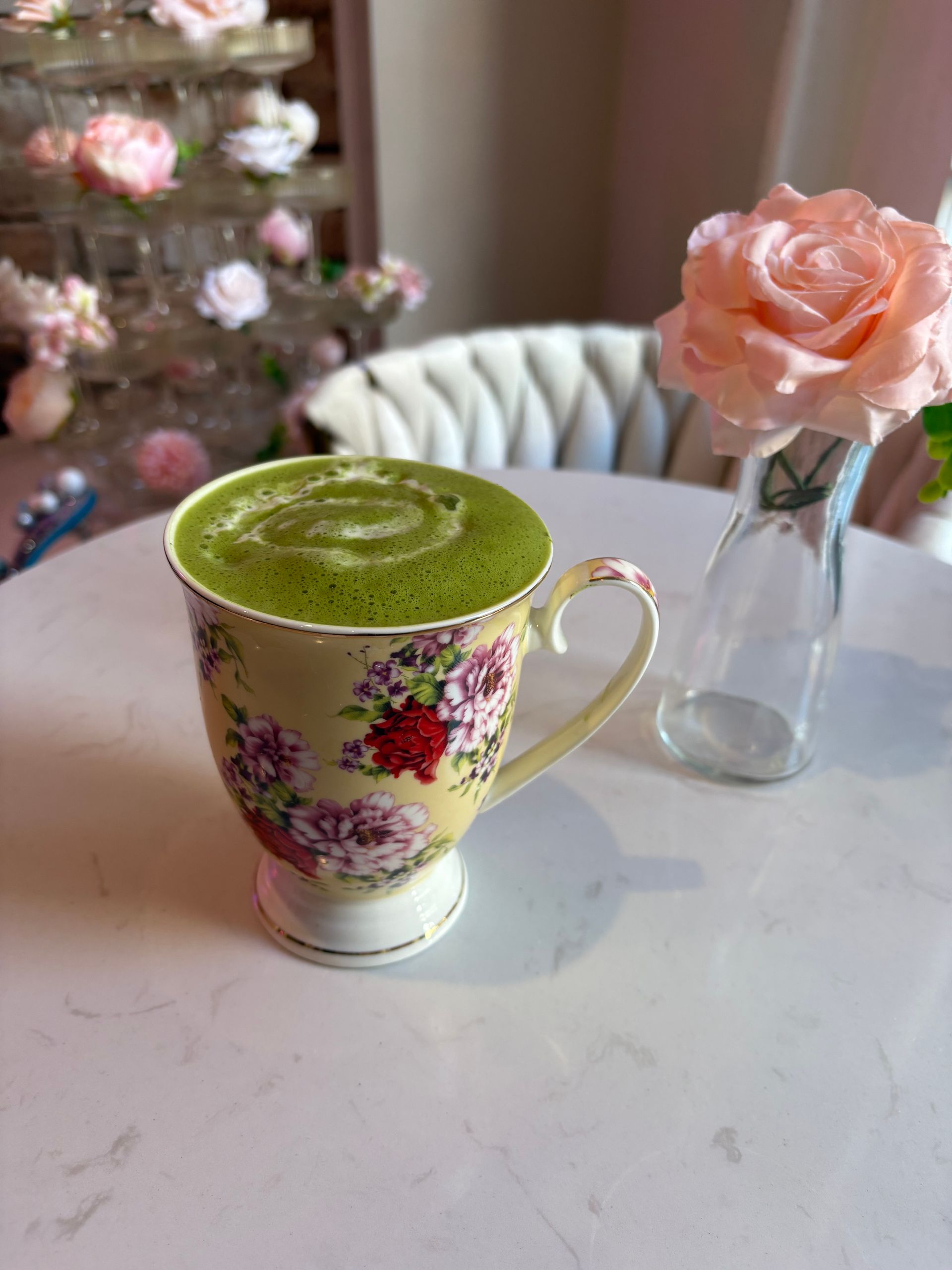 Matcha latte in floral teacup on a white table with a rose in a vase.