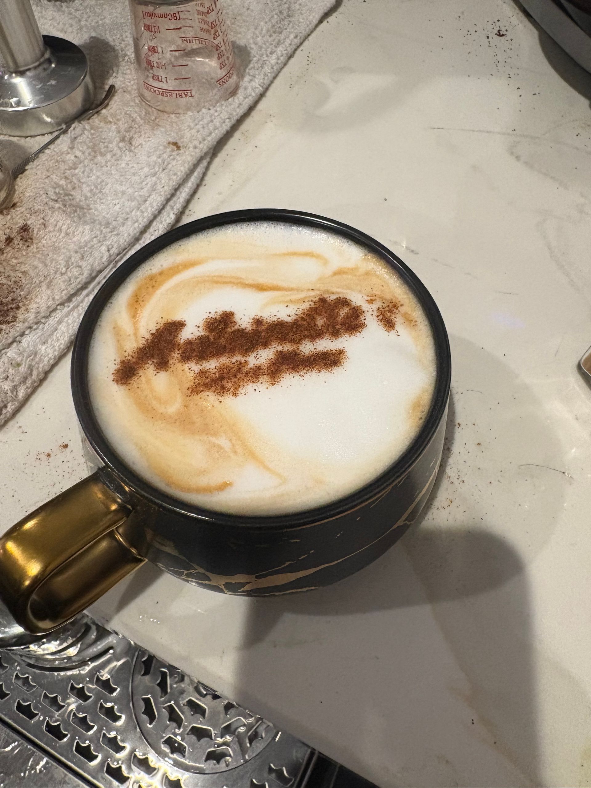 Cappuccino in a black mug with gold handle, cinnamon art on white foam, on a white counter.