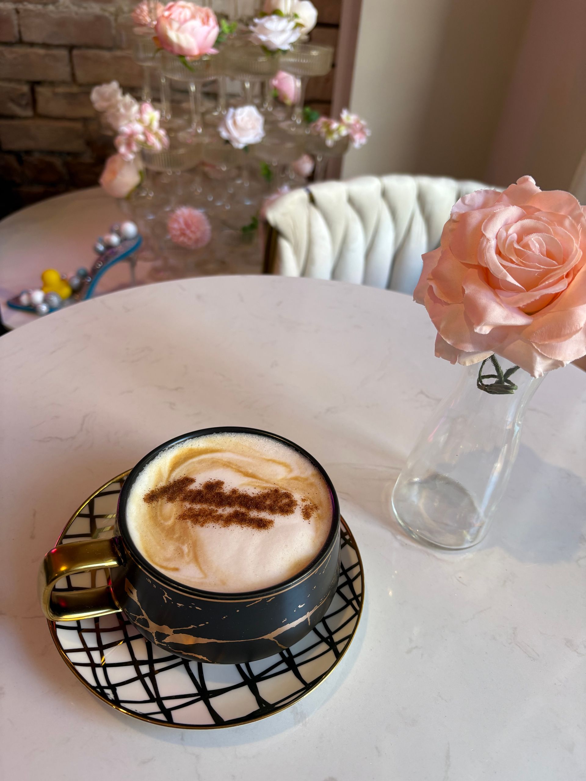 Cappuccino in black and gold cup, single pink rose, and decorative floral arrangement on a white table.