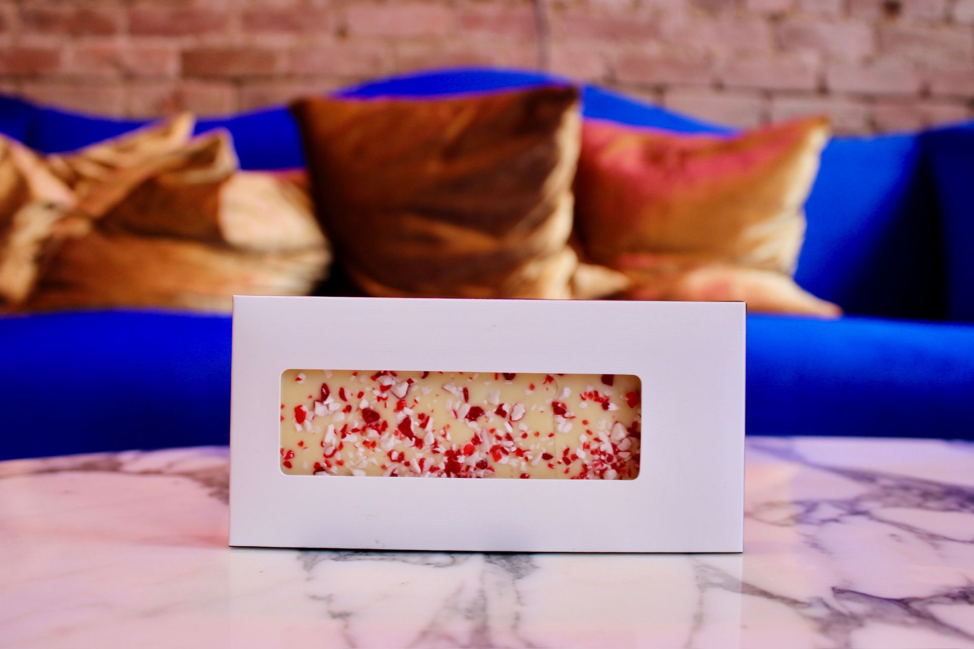 Candy bar with red sprinkles in white box on marble table, blue couch in background.