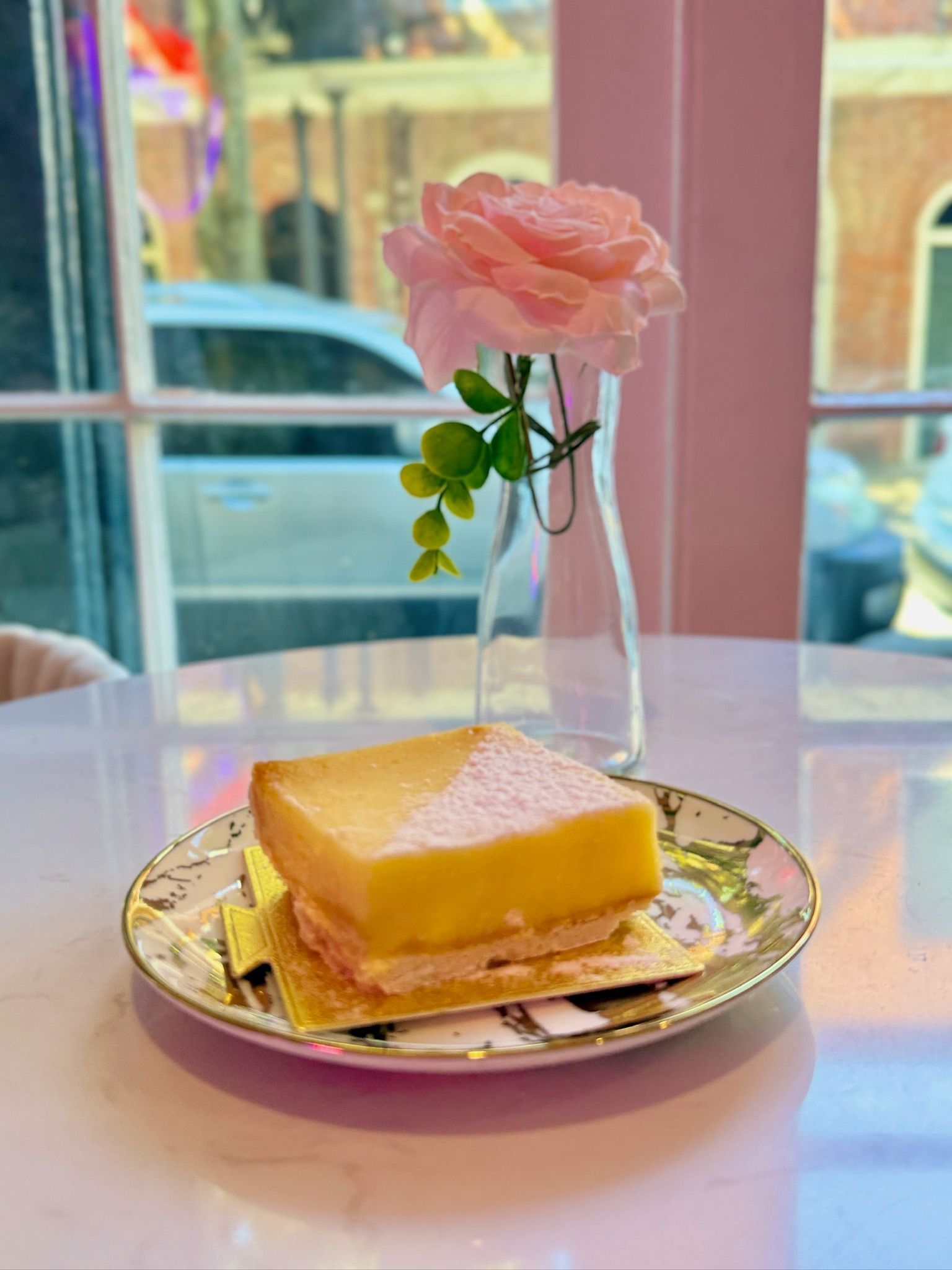 Lemon cake slice on a decorative plate next to a pink rose in a glass vase, on a cafe table.