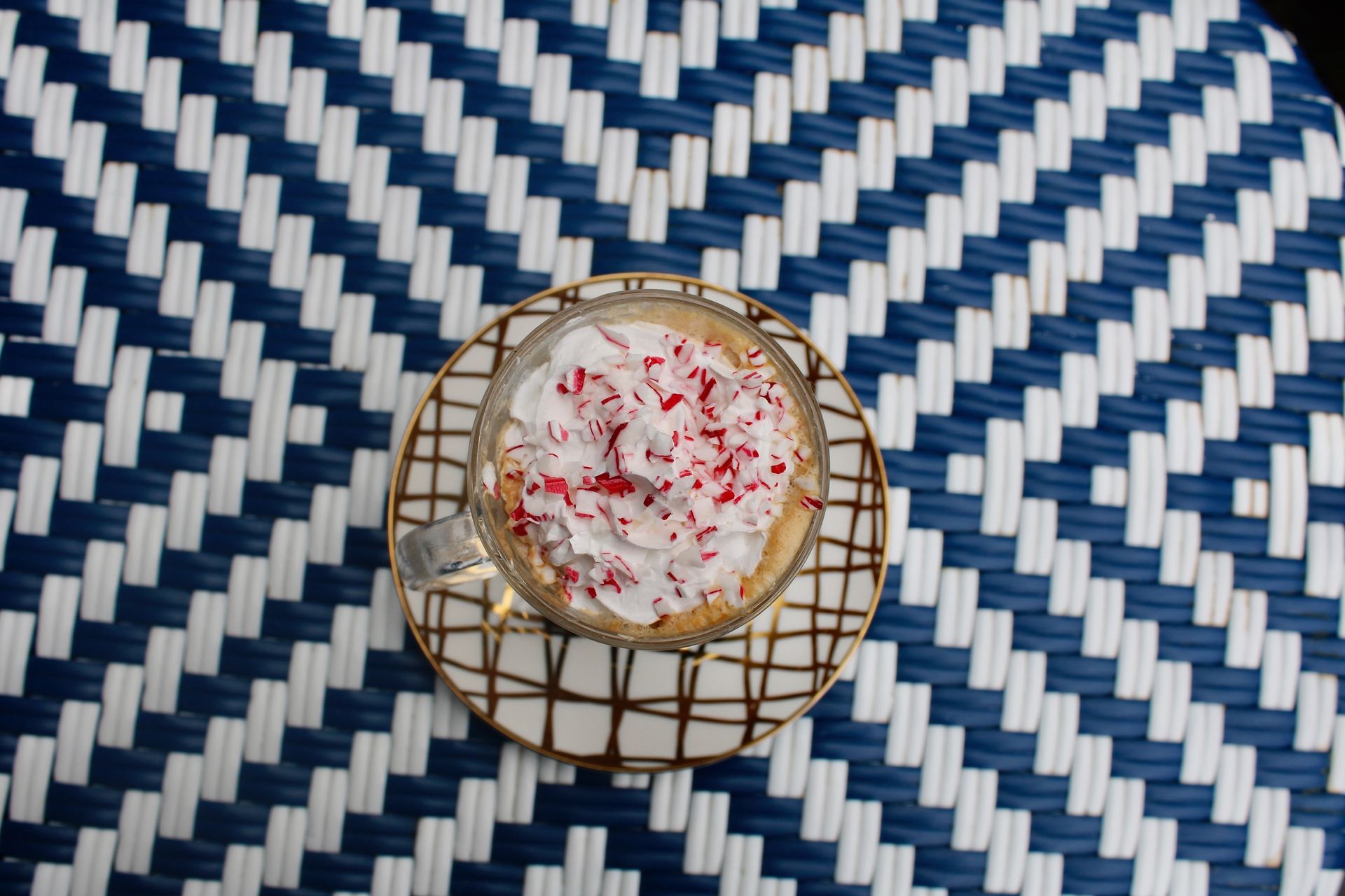 Coffee drink with whipped cream and red sprinkles on a patterned coaster, on a blue and white woven mat.