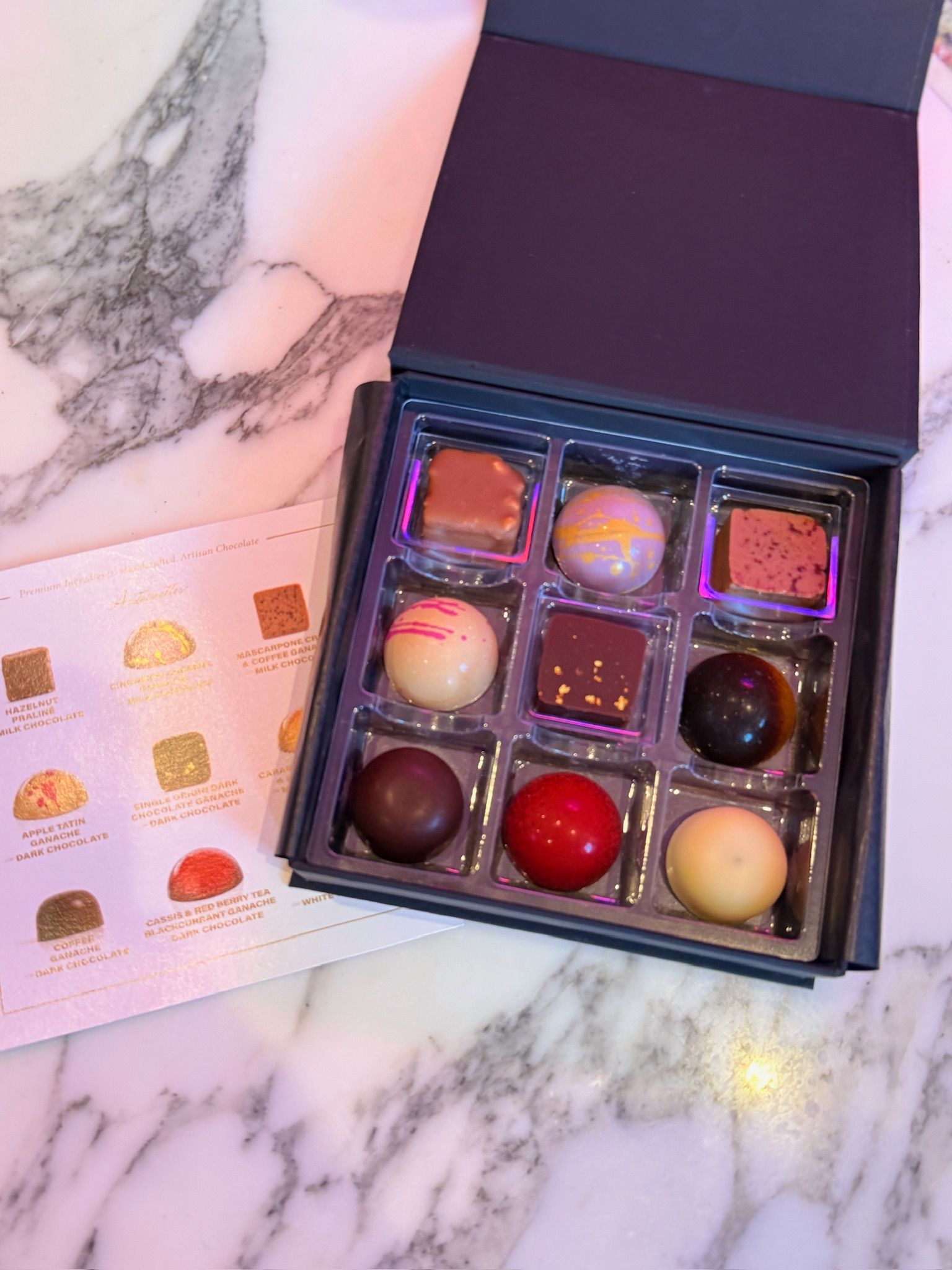 Box of gourmet chocolates in an open blue box, with a flavor key on the left, set on a marble surface.