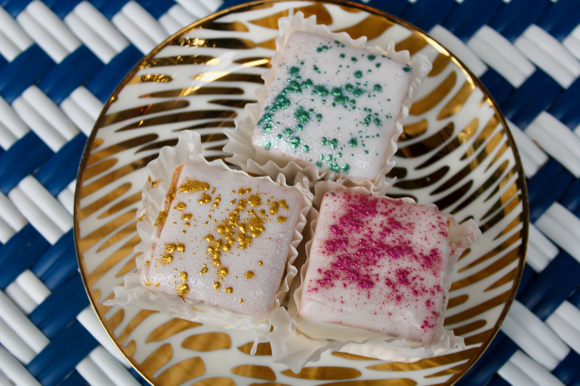 Three pastel-frosted petit fours on a gold-rimmed plate, each with different colored sprinkles, set on a blue and white mat.
