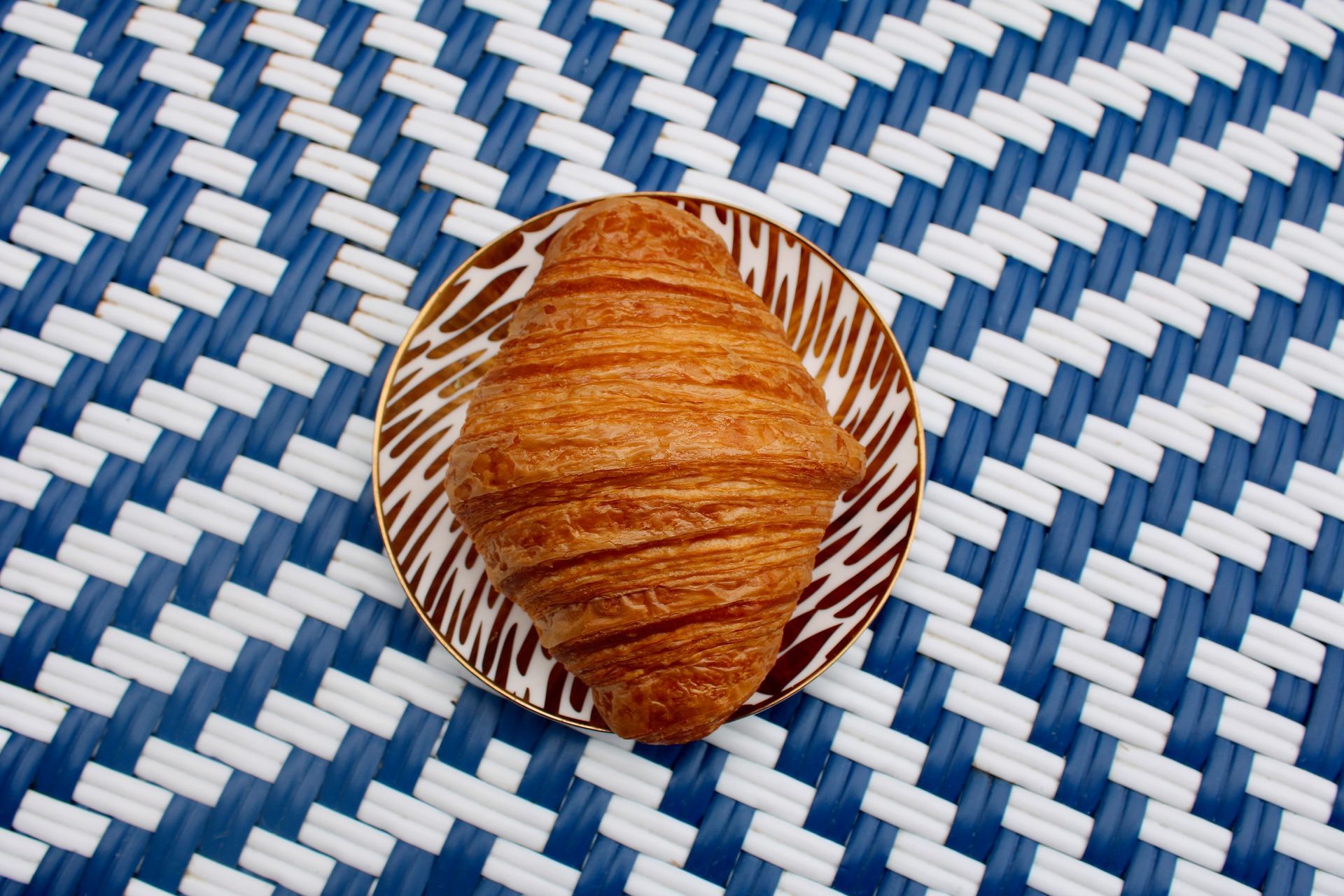 Croissant on a small plate with brown swirls, set on a blue and white woven surface.