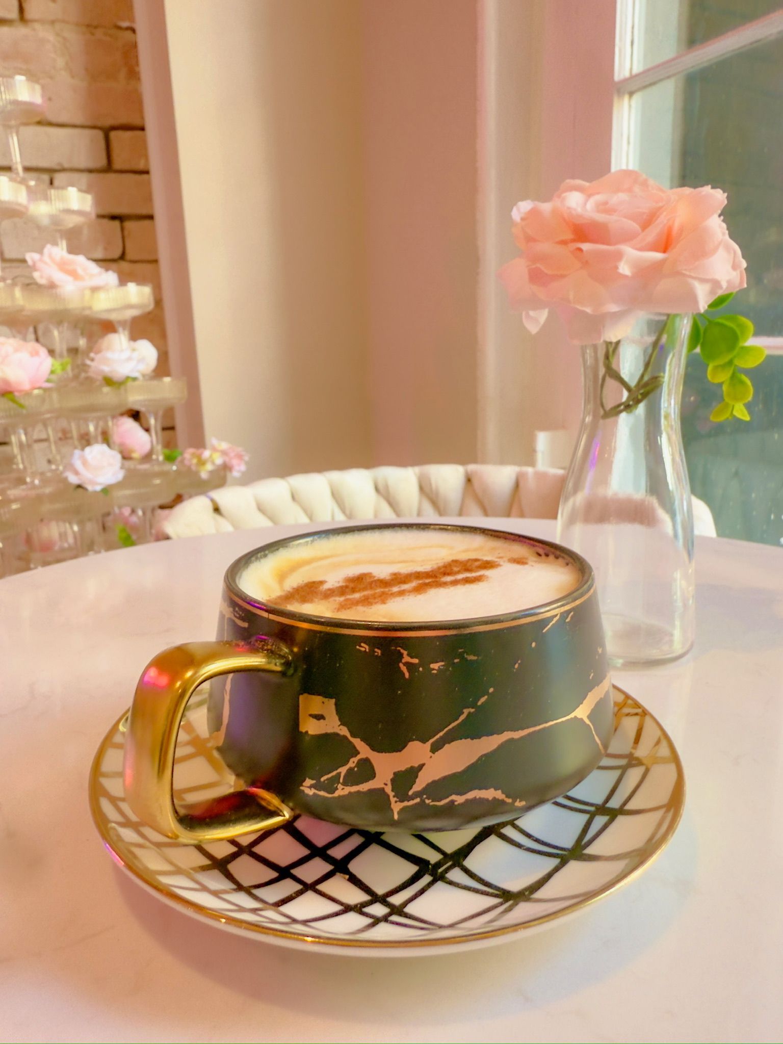 Cup of coffee on a saucer with a rose in a vase on a table, next to a tower of pastries.