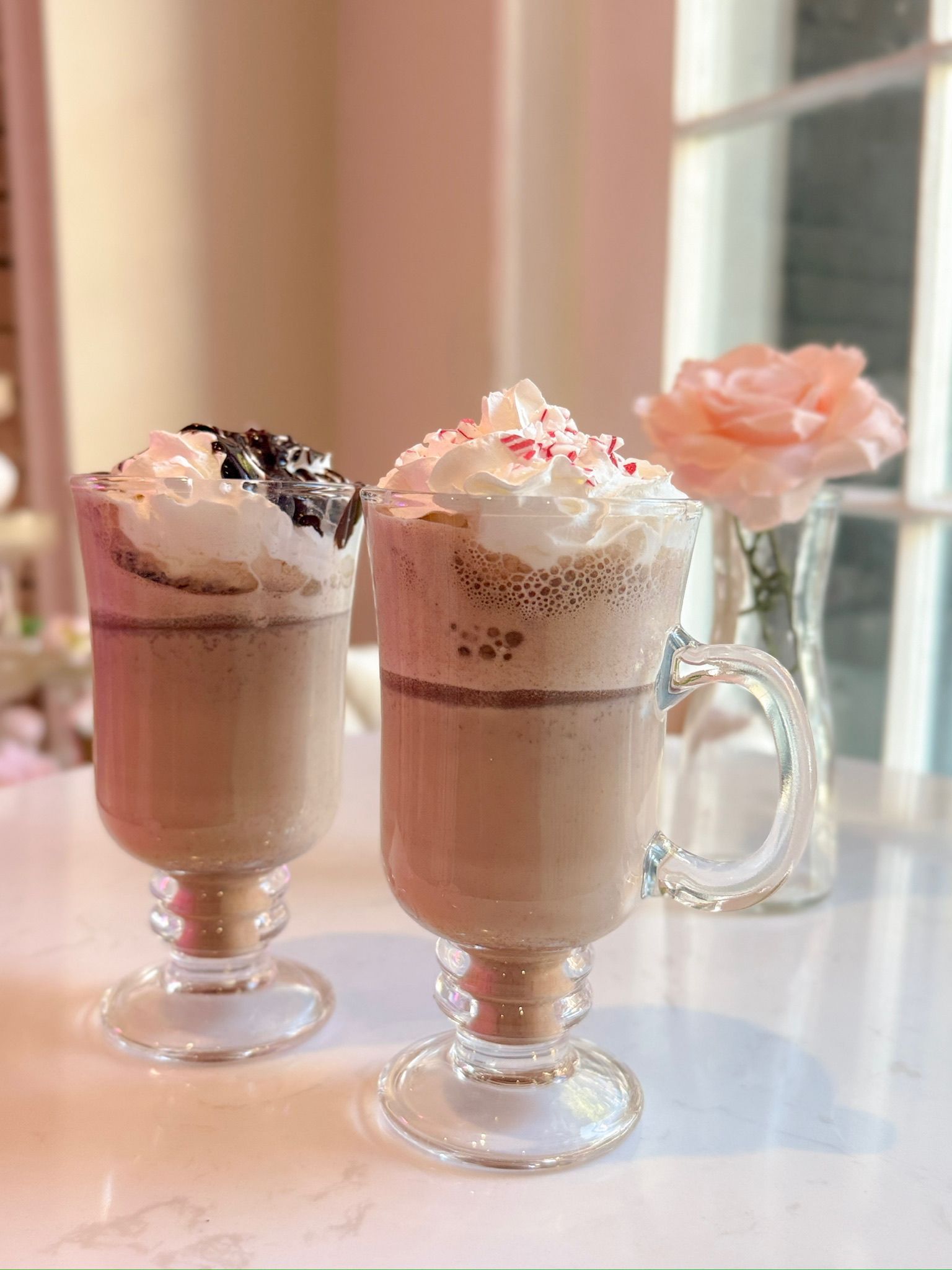 Two frosted coffee drinks with whipped cream, in glass mugs, by a window with a rose.