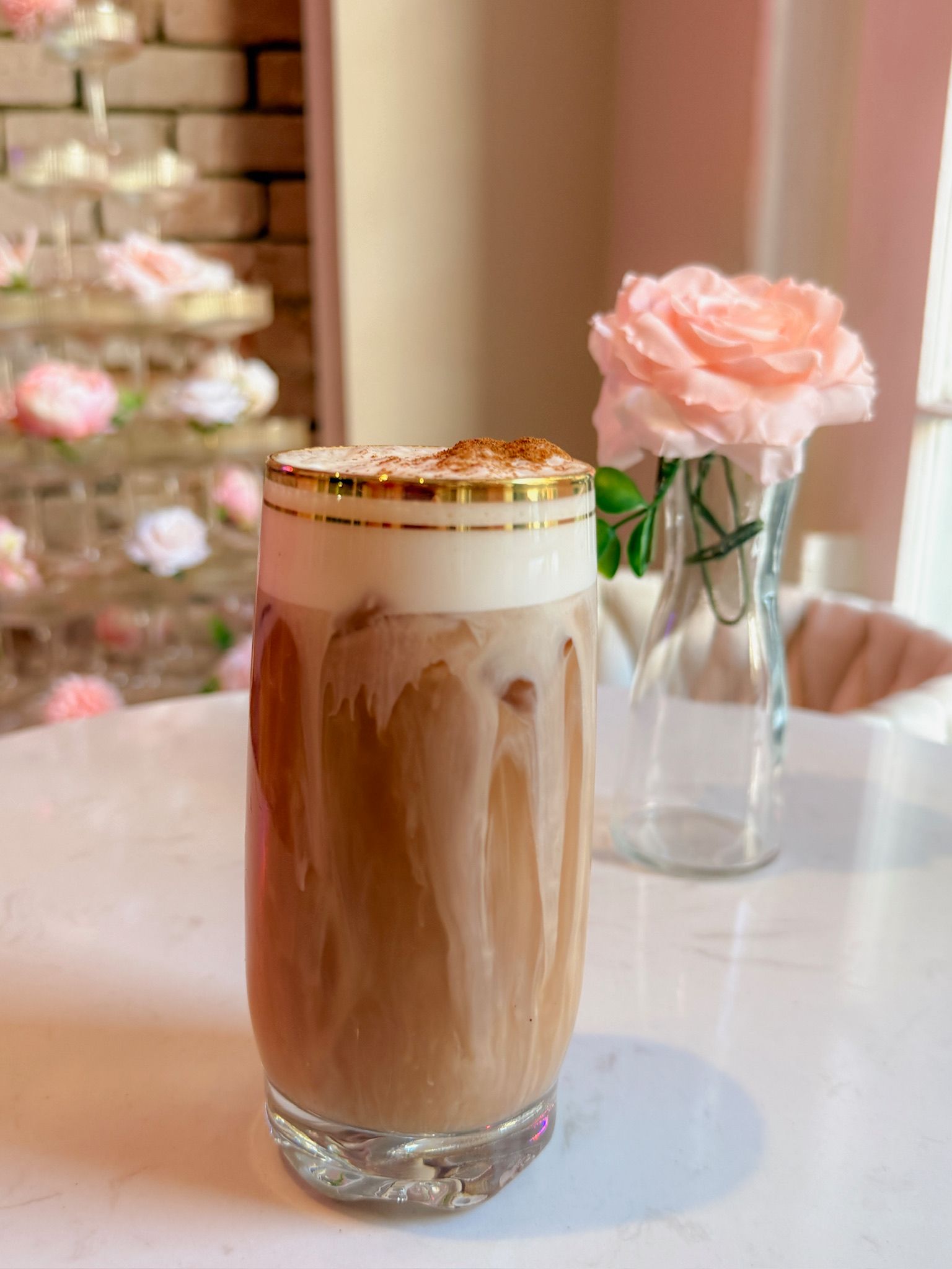 Iced coffee with frothy top, gold-rimmed glass, on a white table. A pink rose and a tiered dessert stand.