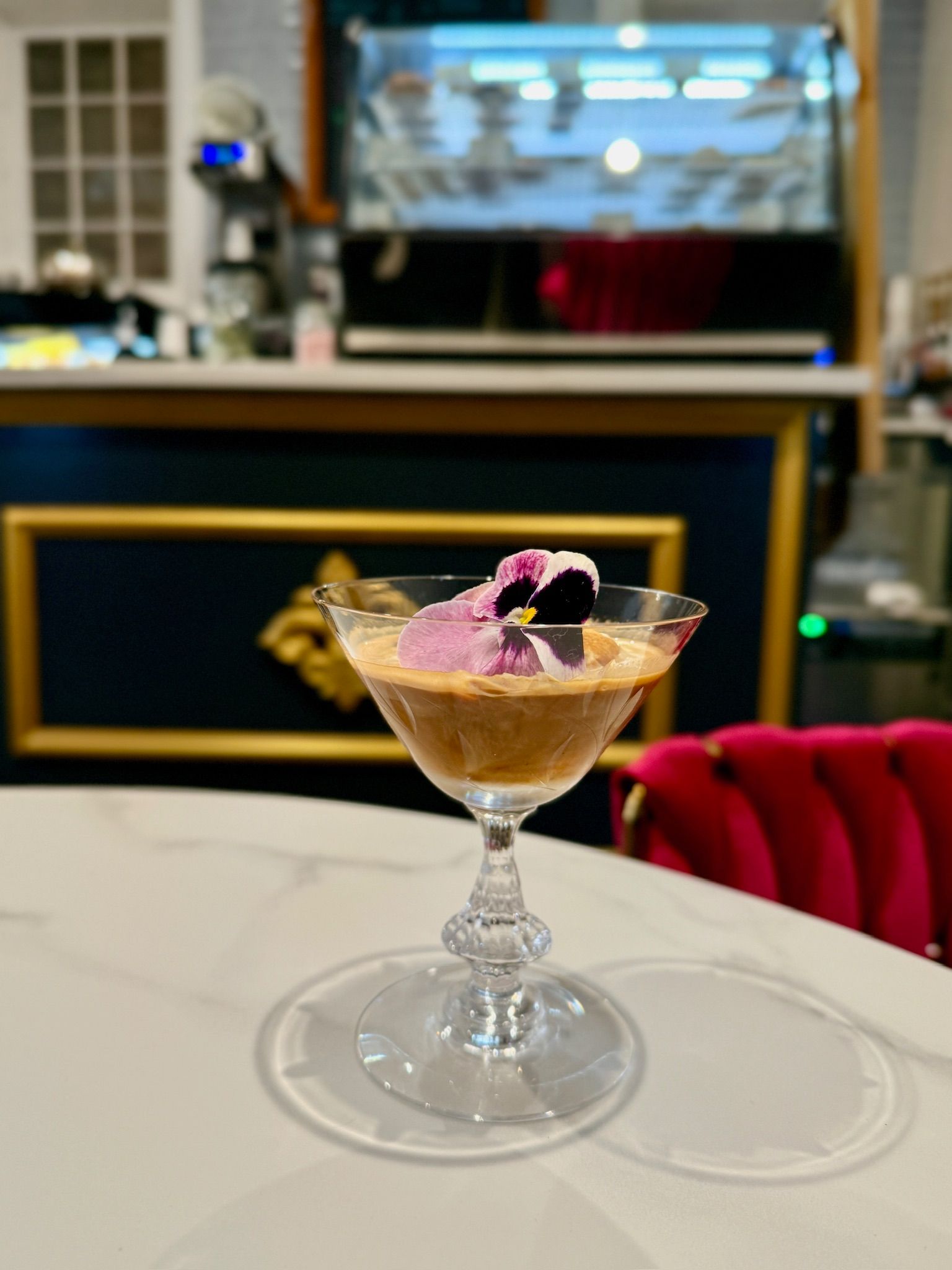 Cocktail in a glass, with a flower garnish, sits on a table. Cafe interior in background.