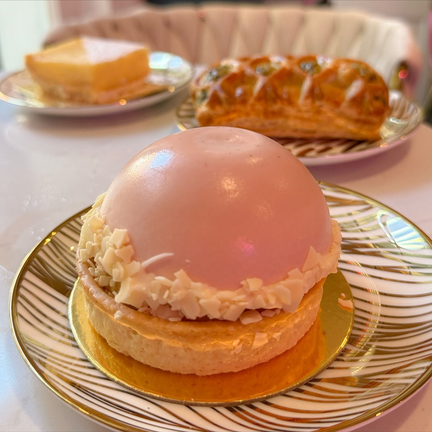Pink-domed pastry on a plate with two other pastries in the background. Desserts are on a white table.