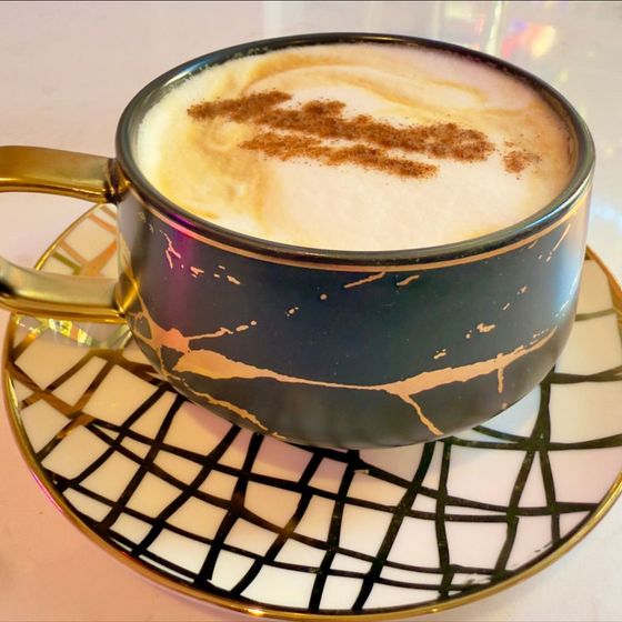 Coffee cup with gold handle on a gold-rimmed saucer with black and white grid pattern. Cinnamon sprinkled on top.