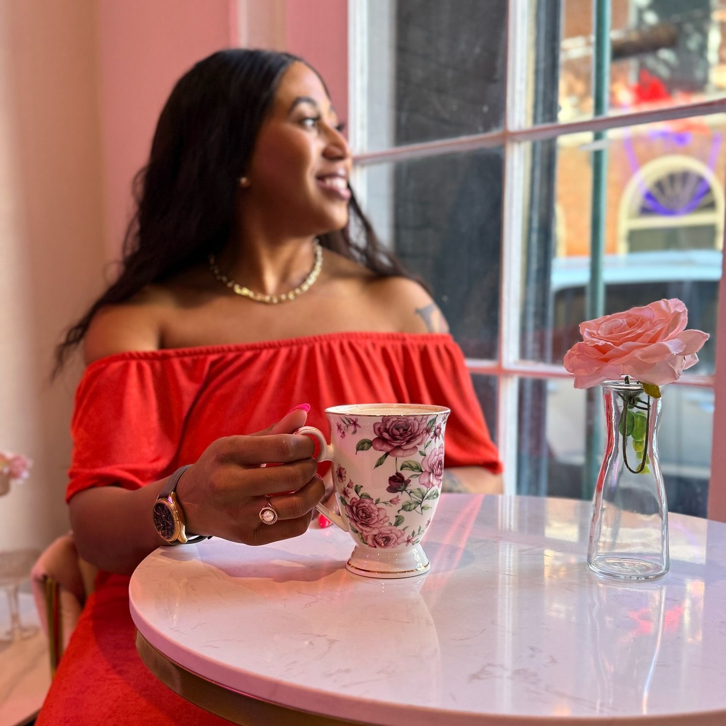 Woman at a table with a floral teacup and pink flower, looking out a window, wearing a red off-the-shoulder top.