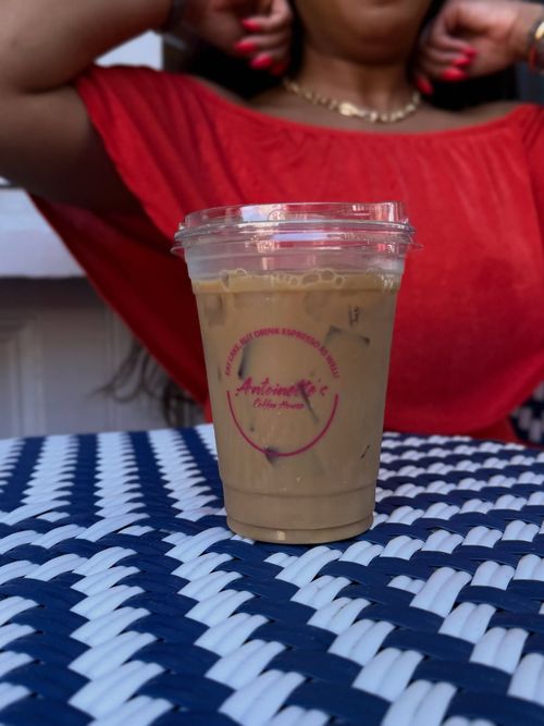 Iced coffee in a clear plastic cup on a blue and white checkered table; woman in red shirt in background.