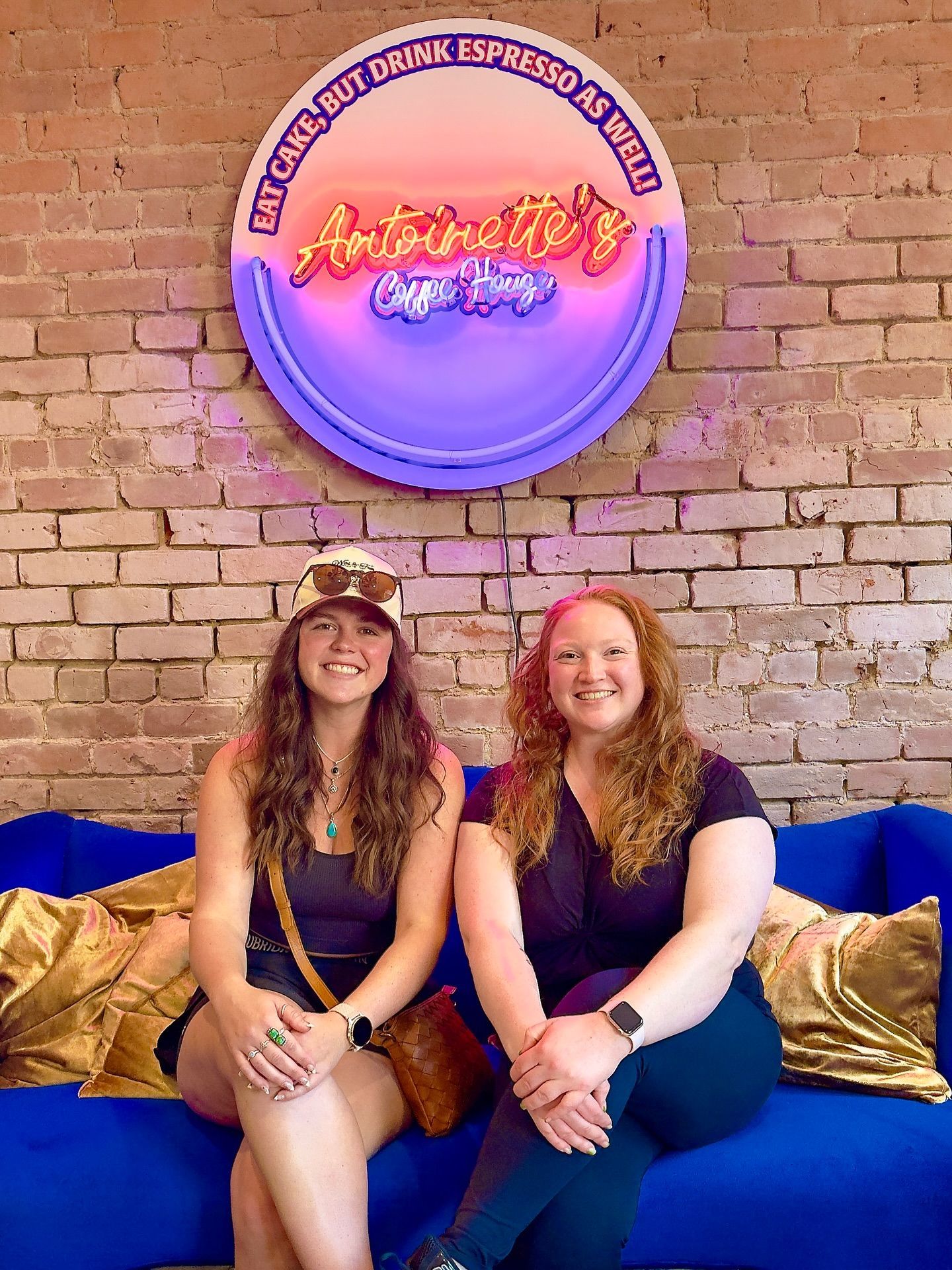 Two smiling women seated on a blue couch in front of a brick wall and neon sign.