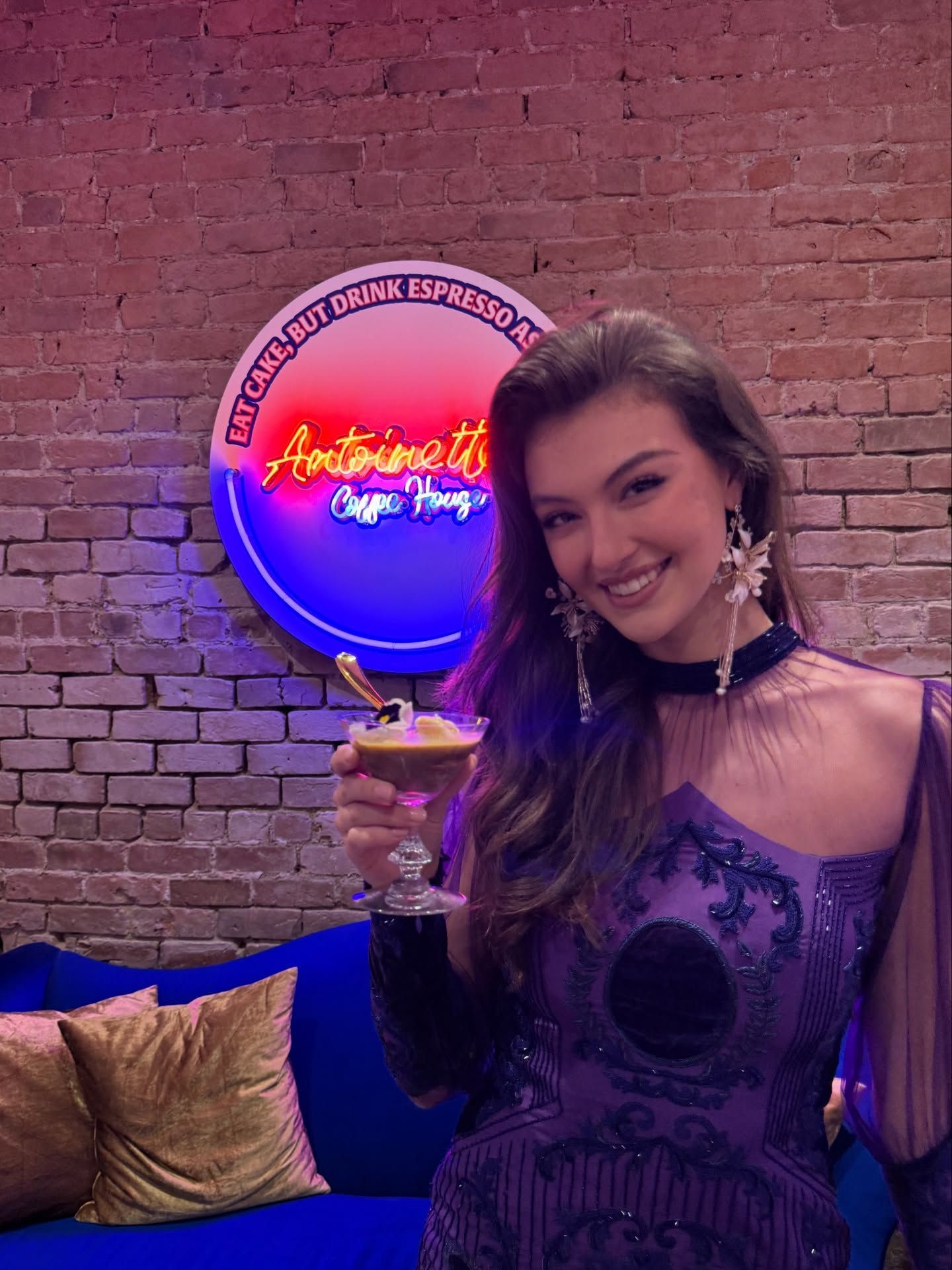 Woman in purple dress, holding a drink, smiles in front of a neon sign.