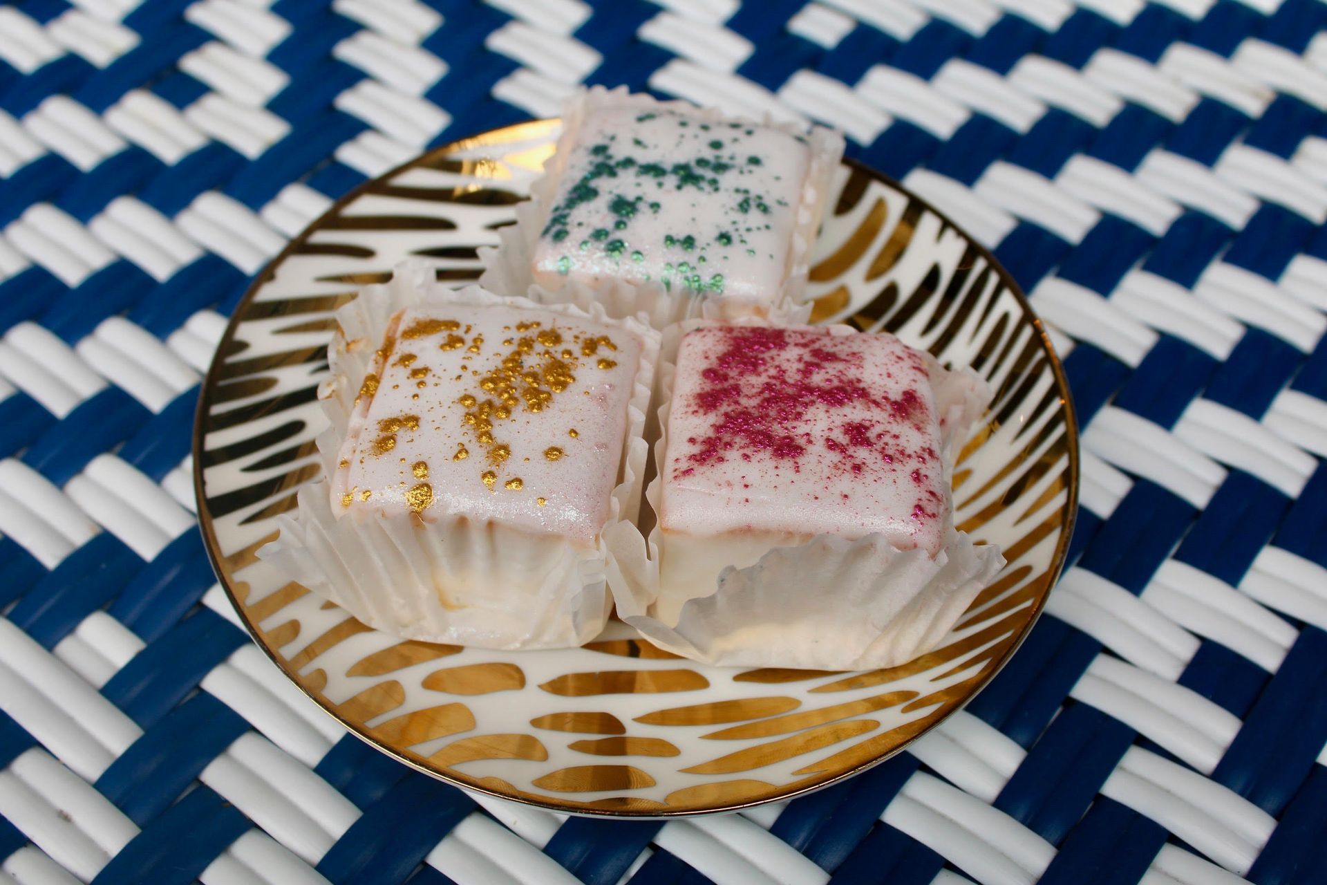 Three square pastries with colored sugar on a gold-rimmed plate, placed on a blue and white woven mat.