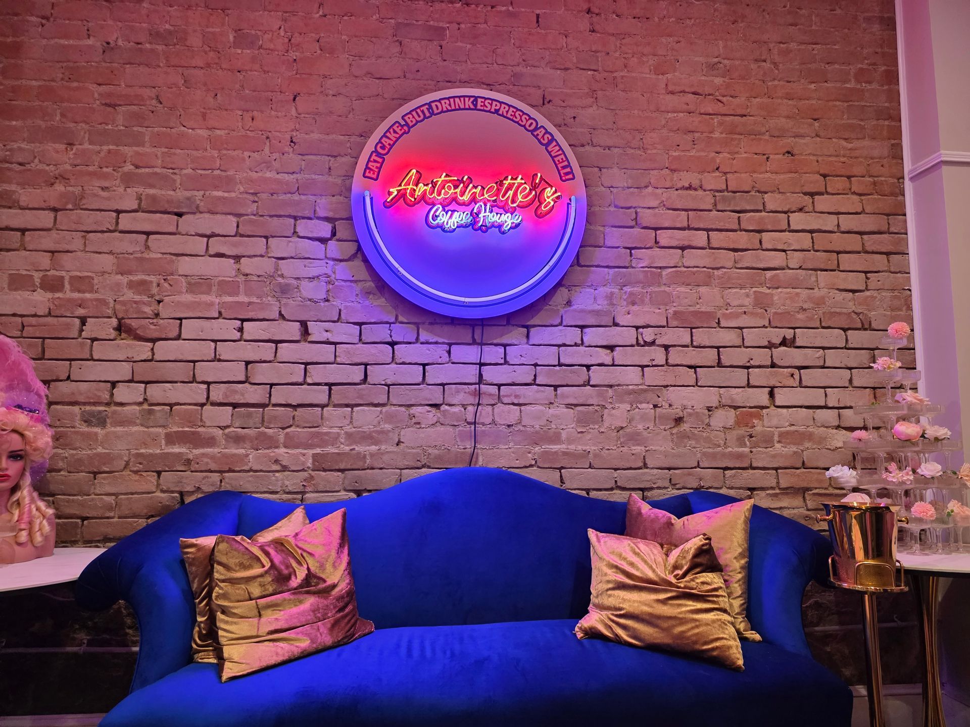 A blue velvet couch with gold pillows sits below a neon sign on a brick wall.