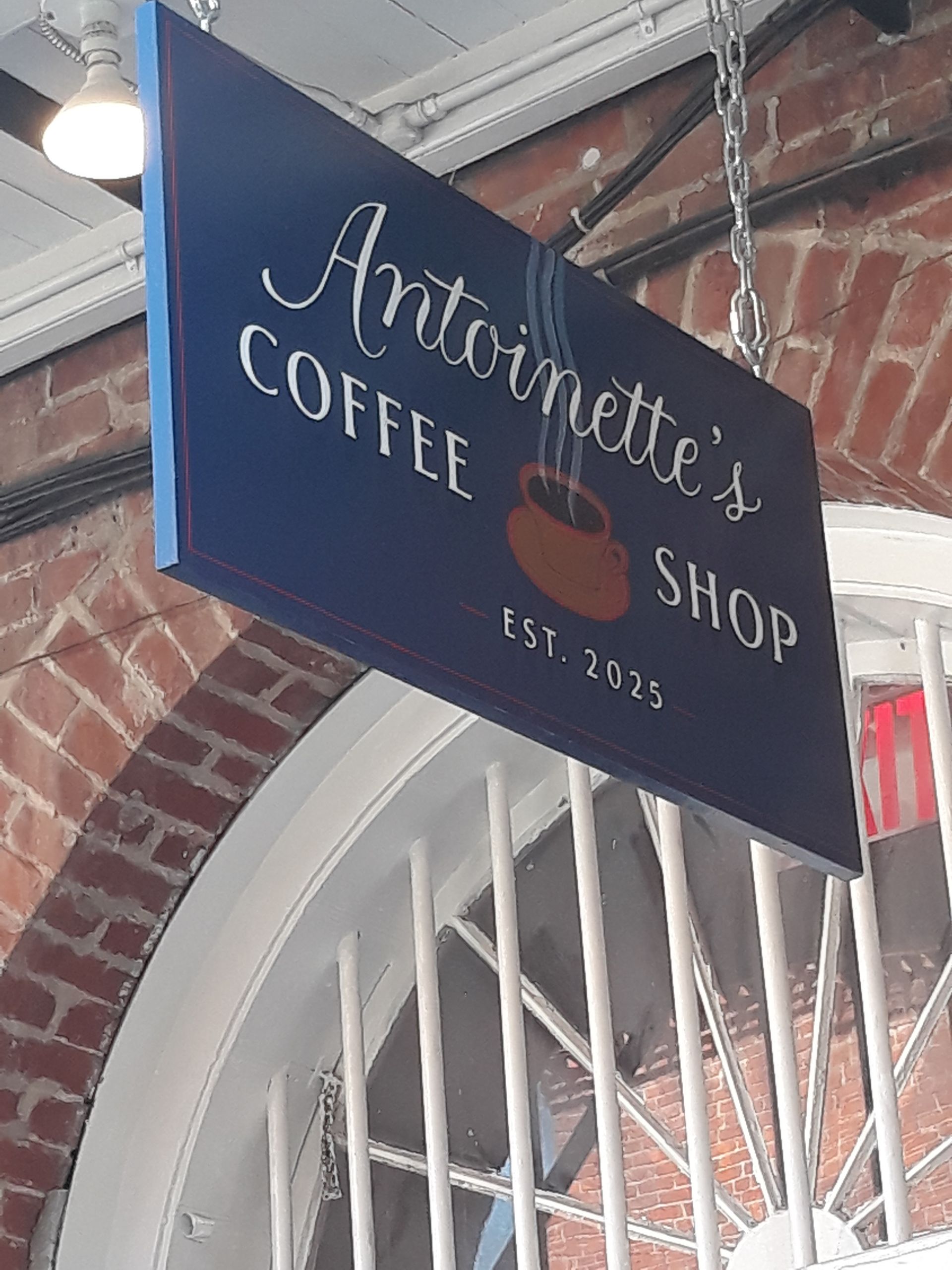 Sign for Antoinette's Coffee Shop, navy blue with white script and a coffee cup graphic, hanging by chains. Est. 1925.