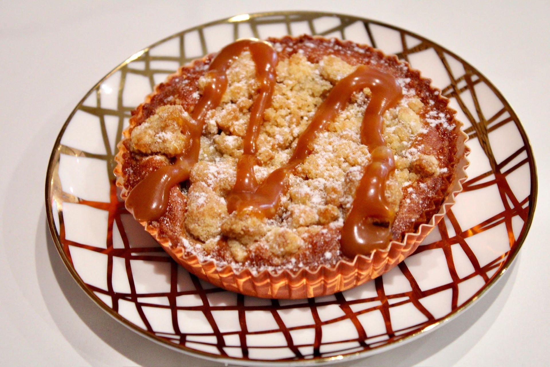 Apple crumble tart on a decorative white and gold plate, drizzled with caramel sauce.